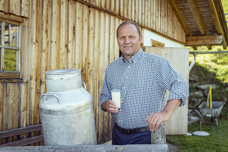 Josef Geisler steht vor einer Hütte und neben einer Milchkanne und hält ein Glas mit Milch in der Hand