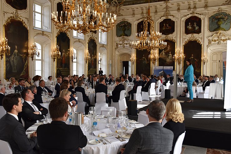 Der Riesensaal in der Hofburg in Innsbruck: Ein großer Raum mit hoger Decke; Goldleuchter hängen von der Decke; an der Wände sind Portraits und Goldverzirrungen; im Saal sind runde weiße Tische aufgestelt um die Menschen sitzen. Am rechten Rand des Fotos ist eine Bühne zu sehen. Auf ihr steht Mair und hält ihre Rede