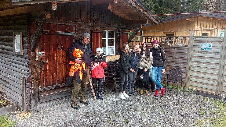 Andreas Waldhauser von der Bezirksforstinspektion Reutte (erster von links) steht mit fünf Schülerinnen der Mittelschulen Untermarkt und Lechtal vor einer Holzhütte mit einer Tür und einem Fenster mit weißem Rahmen. 