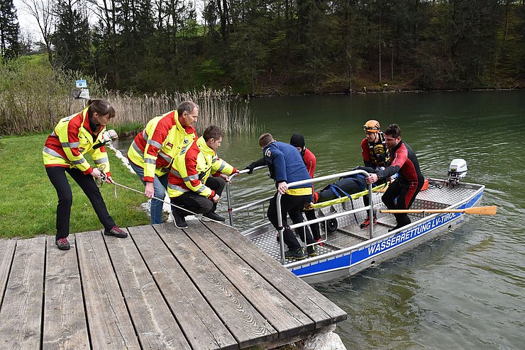 Das Boot der Wasserrettung ist am Steg angekommen: 4 Männer mit Neoprenanzügen stehen im Boot und halten eine Trage. Auf dieser liegt die Verunglückte. Am Steg stehen drei Mitglieder der Wasserettung mit Gelb-Roten Jacken. Sie halten das Boot fest und nehmen die Verunglückte auf der Trage entgegen.