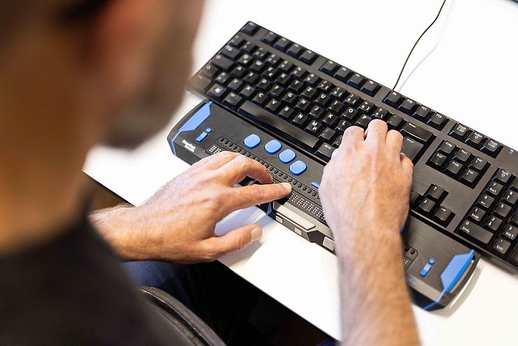 Hände bedienen eine Braillezeile, dahinter eine reguläre Bildschirmtastatur