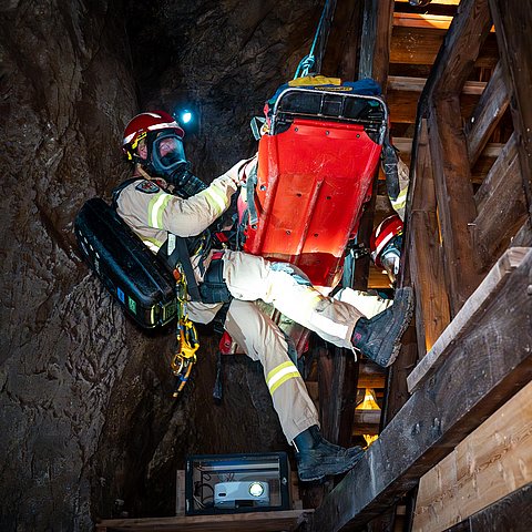 Grubenretter mit Atemschutzmaske und Patient in Trage werden in Stollen abgeseilt.