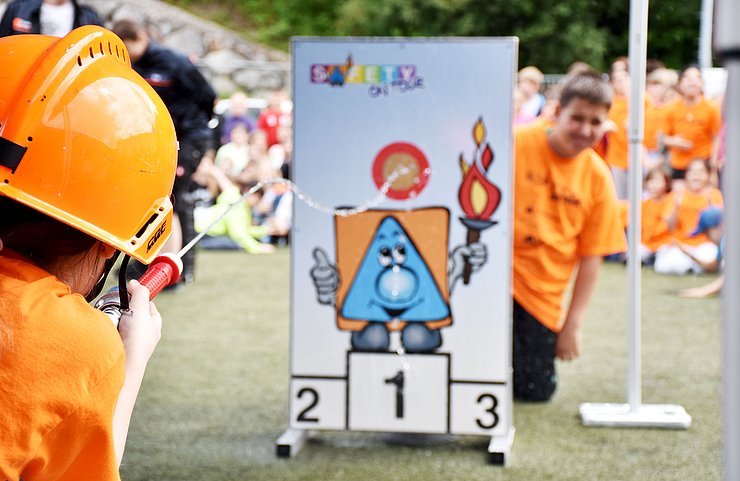 Ein Bub zielt mit dem Schlauch auf die Zieltafel, dahinter schaut ein anderer Schüler hervor.