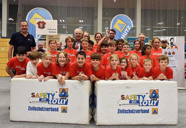 Foto Siegerehrung: Die Kinder der 4mi der Volksschule Kufstein-Zell stehen in roten T-Shirts in zwei Reihen hinter einem weißen Podest und freuen sich über den dritten Platz. 