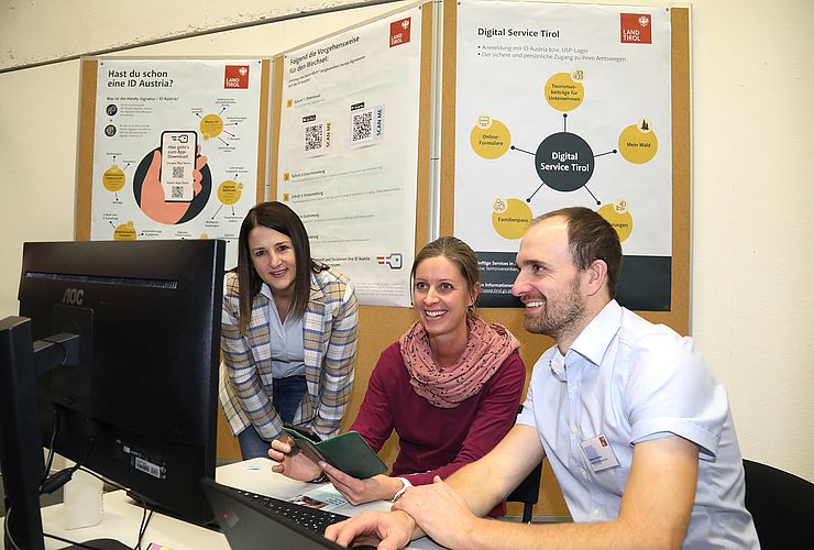 Drei Personen sitzen vor Computer, im Hintergrund Plakate zur ID Austria