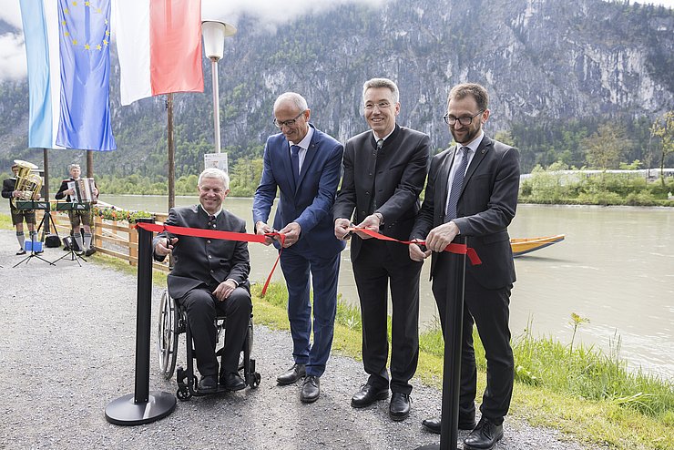 Gruber (im Rollstuhl), Mattle, Lederer und Kolland hinter einem roten Band mit Scheren in der Hand und schneiden das Band durch; im Hintergrund der Inn; im linken Bildrand Fahnen