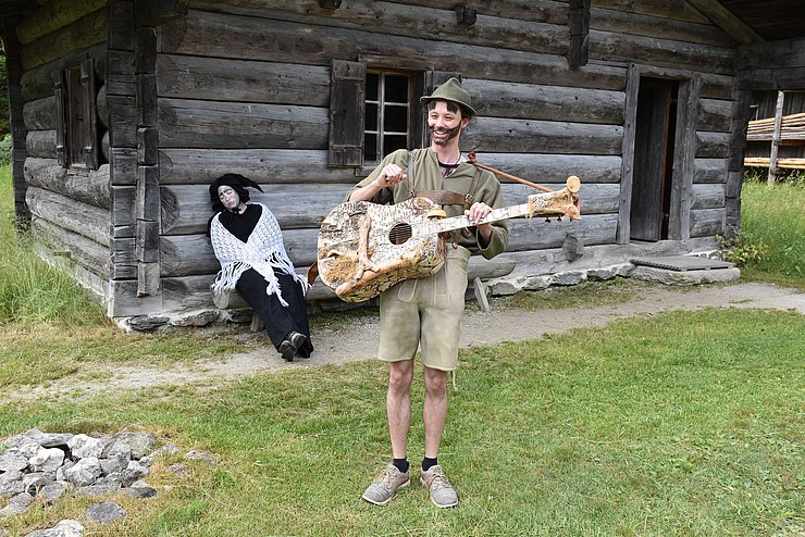 Almajuri hat einen Hut auf und trägt einen Bart. Er spielt auf einer Gitarre vor einer Hütte. Hinter ihm sitzt ein Fabelwesen auf einer Bank vor der Hütte.