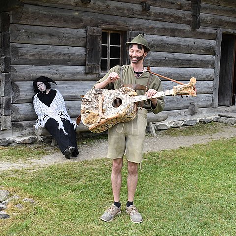 Almajuri hat einen Hut auf und trägt einen Bart. Er spielt auf einer Gitarre vor einer Hütte. Hinter ihm sitzt ein Fabelwesen auf einer Bank vor der Hütte.