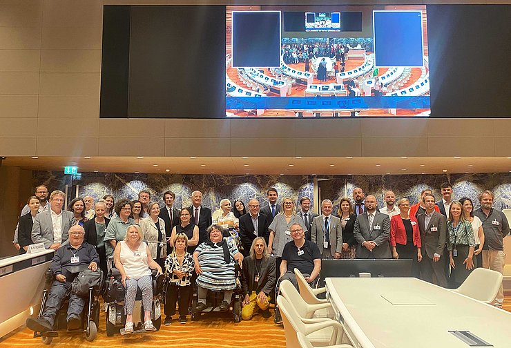 Gruppenfoto, dahinter Bildschirm, der Geschehen aus dem Saal überträgt