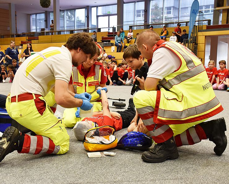 Zwei Rettungssanitäter des Samariterbundes in rot-weiß-neongrüner Uniform knien um ein Kind und und verarzten es. Die Szene ist jedoch nur gespielt.