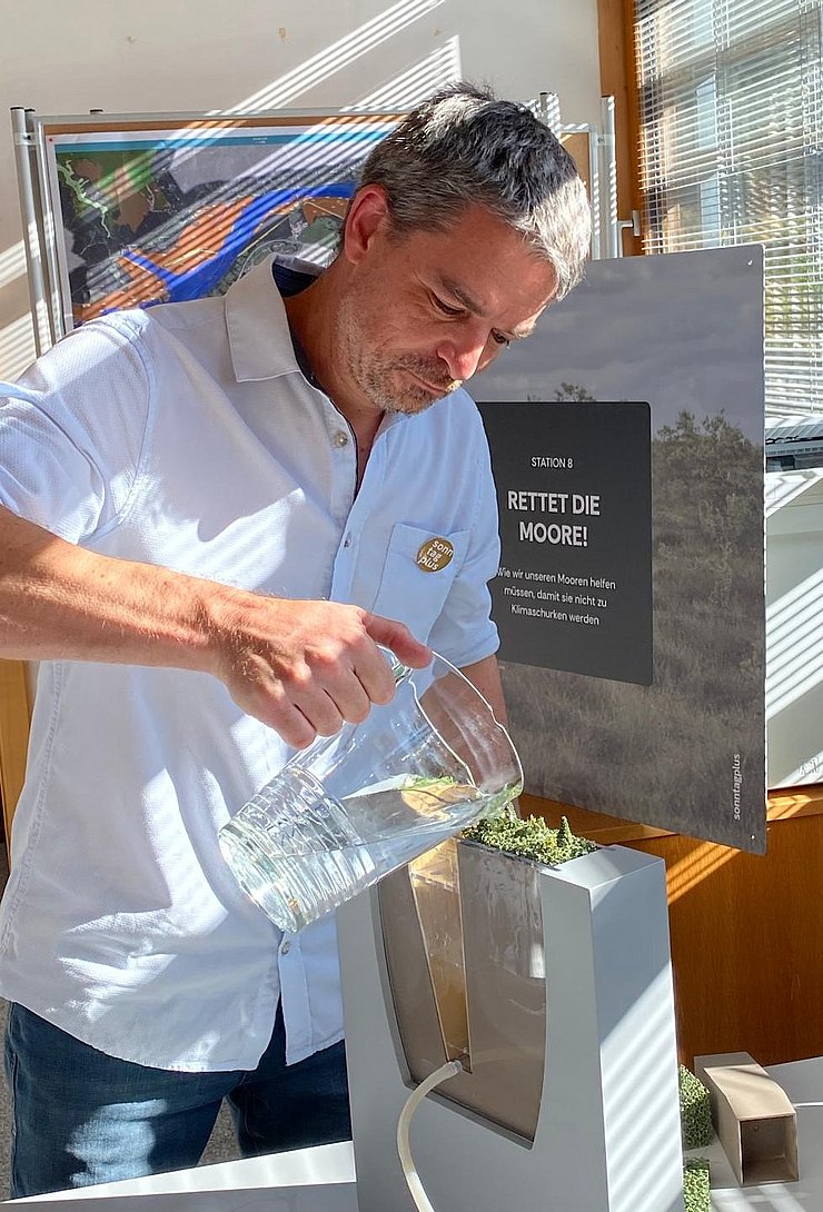 Ein Mann steht an einem Tisch und gießt Wasser aus einer Glaskanne in ein rechteckiges, transparentes Modell mit Pflanzen. Hinter ihm steht eine Tafel mit der Aufschrift „Rettet die Moore!“.