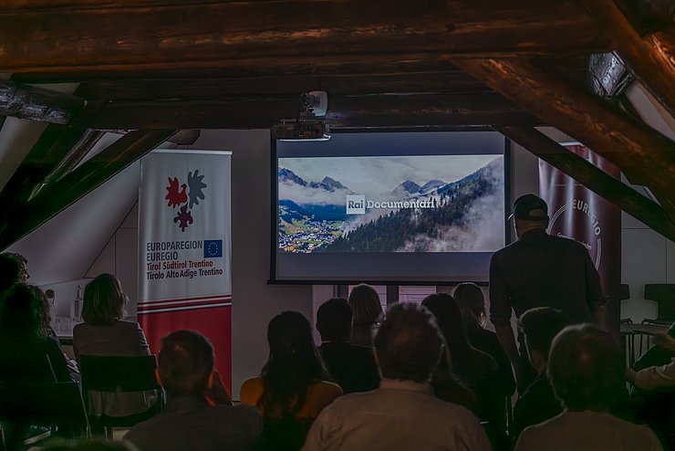 Mehrere Menschen sitzen in einem Saal und schauen auf eine Leinwand.
